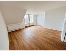 Leerer, heller Wohnraum mit Holzfußboden, weißen Wänden und einer Dachschräge. Große Balkontür-Fensterfront mit Geländerblick sowie ein Heizkörper an der linken Wand.