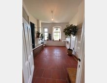 Geräumiges Badezimmer mit roten Fliesen, einer Badewanne auf der linken Seite, großen Fenstern mit Pflanzen auf den Fensterbänken und freistehendem Waschbecken rechts. Ein Topfbaum steht neben einer weiß gefliesten Wand mit Handtuchheizung.