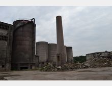 Industriegelände mit verlassenen, verwitterten Gebäuden, darunter mehrere große Silos und ein hoher gemauerter Schornstein. Im Vordergrund sind Trümmer und zerfallene Bauwerke auf einem Betonboden zu sehen, im Hintergrund bewaldete Hügel unter bewölktem Himmel.