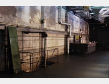 Industriehalle mit verwitterten Wänden, sichtbaren Rohren und Metallstrukturen an der Decke. Rechts im Bild steht eine Bar mit Flaschen auf der Theke, links führt eine Metalltreppe nach unten.