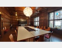 Ein Konferenzraum mit holzvertäfelten Wänden und großen Buntglasfenstern. Eine U-förmige Tischanordnung mit Stühlen umgibt die Mitte des Raumes, darüber hängt eine große, moderne Deckenleuchte.