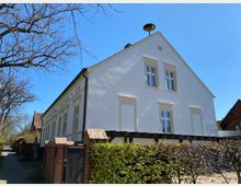 Weiß gestrichenes zweigeschossiges Gebäude mit Satteldach und symmetrisch angeordneten Fenstern im traditionellen Stil. Im Vordergrund stehen eine Hecke, eine Backsteinmauer und eine schmale Gehwegstraße, umgeben von kahlen Bäumen und anderen Gebäuden.