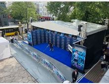 Außenbereich einer Veranstaltungsbühne mit blauem Teppich und Werbebannern für eine Prime-Video-Produktion. Die Bühne ist teilweise überdacht, umgeben von Absperrgittern, und im Hintergrund sind Bäume, ein gelber Bus und Gebäude sichtbar.