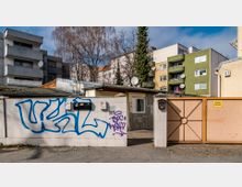 Mehrfamilienhäuser mit Balkonen im Hintergrund, im Vordergrund eine niedrige Mauer mit Graffiti und schwarzem Briefkasten sowie ein metallisches Tor mit einem gelben Schild "Ausfahrt freihalten".