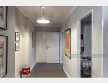 Flur in einem Wohngebäude mit hellgrauen Wänden und einer weißen Decke, beleuchtet durch eine zentrale Hängelampe. An den Wänden hängen Bilder und Poster, ein weißes Türpaar ist am Ende des Flurs zu sehen, und rechts führt eine Tür in einen Raum mit Tisch und Stuhl.