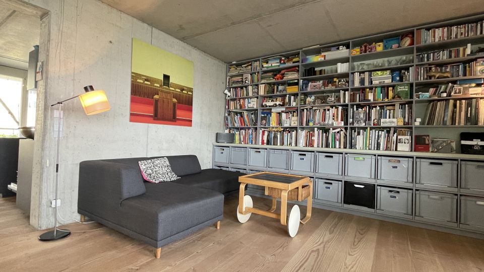 Wohnzimmer mit moderner Einrichtung, bestehend aus einem grauen Sofa, einem Holz-Tisch auf Rädern, einer Stehlampe und einer großen, wandfüllenden Regalwand mit Büchern und Aufbewahrungsboxen. Die Wände sind aus Sichtbeton, der Boden ist aus hellem Holz.