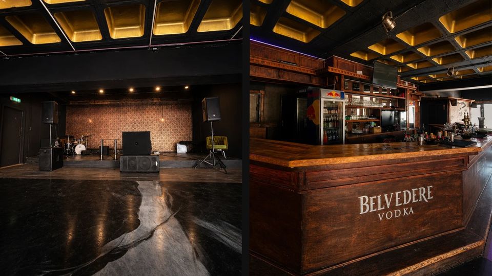 Innenraum eines Musikclubs mit kleiner Bühne, Lautsprechern und Schlagzeug vor gemusterter Rückwand sowie einer Decke mit goldfarbenen Kassetten. Daneben eine lange Bar aus dunklem Holz mit Flaschenregalen und dem Schriftzug „Belvedere Vodka“ an der Front.