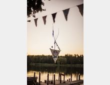 Ein hölzerner Steg an einem ruhigen See bei Sonnenuntergang, mit einem dekorativen Stoffbanner und Girlanden an einem Mast. Im Hintergrund sind Bäume und Schilf am Ufer sichtbar.