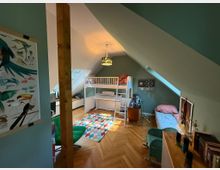 Dachgeschoss-Kinderzimmer mit schrägen Wänden, einem Hochbett, einer niedrigen Fensterbank unter einem Dachfenster und einer bunten Teppichfläche auf Holzboden. Im Raum befinden sich außerdem ein einzelnes Bett, ein kleiner Schreibtisch und verschiedene Dekorationen.