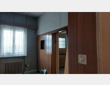 Innenraum eines Altbaus mit einer Wand aus Holzvertäfelung, einer geöffneten Tür und einem Fenster mit Spitzenvorhang. Ein Heizkörper befindet sich unter dem Fenster, und die Wand ist teilweise mit gemusterten Tapeten bedeckt.