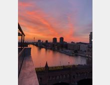 Blick auf die Oberbaumbrücke in Berlin bei Sonnenuntergang, mit der Spree in der Mitte des Bildes und der Skyline der Stadt im Hintergrund, einschließlich des Fernsehturms. Ein gelber Zug fährt über die Brücke, während der Himmel in leuchtenden Orange- und Rosatönen leuchtet.
