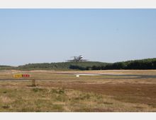 Ein Flugzeug befindet sich im Landeanflug über einer Landebahn, umgeben von einer grasbewachsenen Fläche und einem Wald im Hintergrund. Im Vordergrund sind ein gelbes Richtungs- und Rollwegzeichen sowie asphaltierte Rollwege sichtbar.