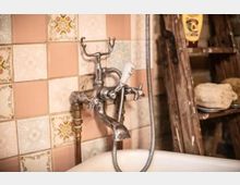 Nahaufnahme eines nostalgischen Wasserhahns mit Handbrause über einer Badewanne, vor einer Wand mit rosafarbenen und gemusterten Fliesen. Rechts im Bild ist eine hölzerne Ablage mit Schwämmen und Pflegeprodukten zu sehen.