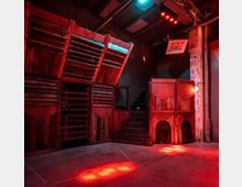 Innenraum eines modernen Veranstaltungsorts mit industriellem und rustikalem Design, geprägt von hölzernen Strukturen, offenen Treppen und Betonwänden. Rotes und blaues Licht beleuchtet den Raum stimmungsvoll.