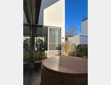 Moderne Terrasse mit minimalistischem Design, umgeben von hohen Glasflächen und weißen Gebäudewänden. Im Vordergrund ein runder Whirlpool mit Abdeckung, daneben eine Holzbank und Pflanzen in Töpfen, im Hintergrund blauer Himmel und kahler Baum.