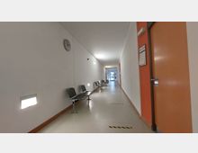 Langer, heller Flur mit weißen Wänden, an denen Stühle aus schwarzem Kunststoff aufgereiht stehen. Auf der rechten Seite befinden sich Türen mit orangenen Paneelen, und am Ende des Flurs ist eine Glastür sichtbar.