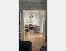 Wohnzimmer in einem hellen, klassischen Stil mit Holzboden und großen Fenstern, die von weißen Vorhängen umrahmt sind. Im Raum befinden sich ein Schreibtisch, ein Rattanstuhl, ein kleiner Teppich sowie eine hängende Holzlampe; eine Blumenkontaktwand ist im Vordergrund sichtbar.