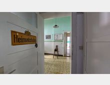 Blick durch eine geöffnete Zimmertür mit dem gelben Schild „Heimatstube“ in einen Raum mit grünen Wänden, einfachen Möbeln und ausgestellten Bildern an der Wand. Im Hintergrund führt eine weitere offene Tür in einen helleren angrenzenden Raum.