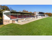 Historische Tribüne eines Sportstadions mit roten Sitzreihen und Überdachung, daneben weitere Gebäude im gleichen Stil. Der Vordergrund zeigt eine große, gepflegte Rasenfläche, umgeben von Bäumen in herbstlicher Farbgebung.