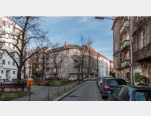 Straßenszene in einer Wohngegend mit mehrstöckigen Wohnhäusern im historischen Stil, Bäumen ohne Laub, parkenden Autos und Gehwegen mit Sitzbänken. Im Hintergrund ragt ein Gebäude mit rotem Ziegeldach hervor.