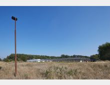Weite, brachliegende Wiese mit vereinzeltem hohen Metallmast im Vordergrund; im Hintergrund mehrere Gewächshäuser aus Glas und Metall sowie eine Baumreihe unter klarem, blauem Himmel.