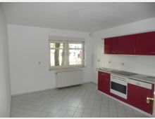 Helle Küche mit weißen Fliesenboden und einer Einbauküche mit roten Schränken, Herd, Backofen und Spüle. Ein Fenster mit Heizkörper darunter zeigt den Blick auf Bäume und eine Außenstraße.