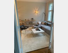 Modernes Wohnzimmer mit heller Wandfarbe und Holzboden. Möbel umfassen ein graues Ecksofa, einen weißen Couchtisch, und einen schwarzen Sessel; an der Wand hängen Regalboards mit Dekoration und Fotos, während eine strahlende Wandleuchte zentral platziert ist.