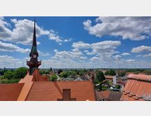 Kirchturm mit spitzem Dach und Uhr in einer ländlichen Kleinstadt, umgeben von roten Ziegeldächern, Bäumen und Windrädern in der Ferne unter einem bewölkten blauen Himmel.