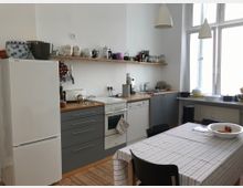 Helle Küche mit einer Kombination aus grauen Unterschränken und einer Holzarbeitsplatte. Ein weißer Kühlschrank steht links, darüber ein Regal mit Geschirr und Küchenutensilien. Rechts befinden sich ein großes Fenster mit viel Tageslicht, ein Esstisch mit gestreifter Tischdecke und Stühlen.