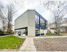 Zweistöckiges, modernes Gebäude aus Sichtbeton mit dunkelblauen Fassadenbändern und großen Fensterflächen, umgeben von kahlen Bäumen. Davor liegen gepflasterte Wege, Rasenflächen und ein Außenbereich mit Treppe und kleinen Terrassenstufen.