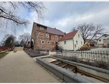 Historisches Fachwerkgebäude mit rotem Ziegeldach und integriertem Wasserrad, umgeben von weiteren Gebäuden und einem gepflasterten Hof mit schmiedeeisernem Zaun; im Vordergrund ein Weg und angelegte Wasserkanäle.