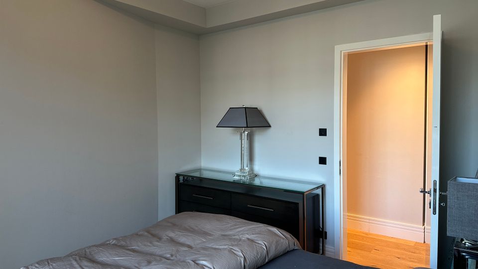 Schlichtes Schlafzimmer mit grauen Wänden und Bett im Vordergrund; an der Wand steht eine schwarze Kommode mit Glasplatte und Tischlampe. Rechts führt eine offene Tür in einen hell beleuchteten Flur mit Holzfußboden.