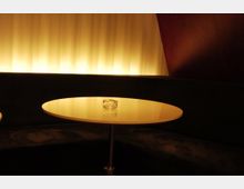 Runder Tisch mit glatter, heller Oberfläche in einem dunklen Raum, beleuchtet von einer weichen, gelblichen Lichtquelle im Hintergrund; auf dem Tisch steht ein Glas-Aschenbecher.