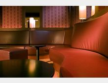 Ein gemütlicher Loungebereich mit geschwungenen roten Polstersofas, kleinen runden Tischen aus dunklem Holz und gedämpfter Beleuchtung durch Stehlampen. Im Hintergrund sind gemusterte Vorhänge und runde Fensteröffnungen zu sehen.