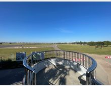 Blick von einer erhöhten Plattform mit Metallgeländer auf eine weitläufige, offene Wiesenlandschaft mit vereinzelten Bäumen und einem asphaltierten Weg. Im Hintergrund sind Gebäude und horizontale Strukturen unter einem klaren blauen Himmel sichtbar.