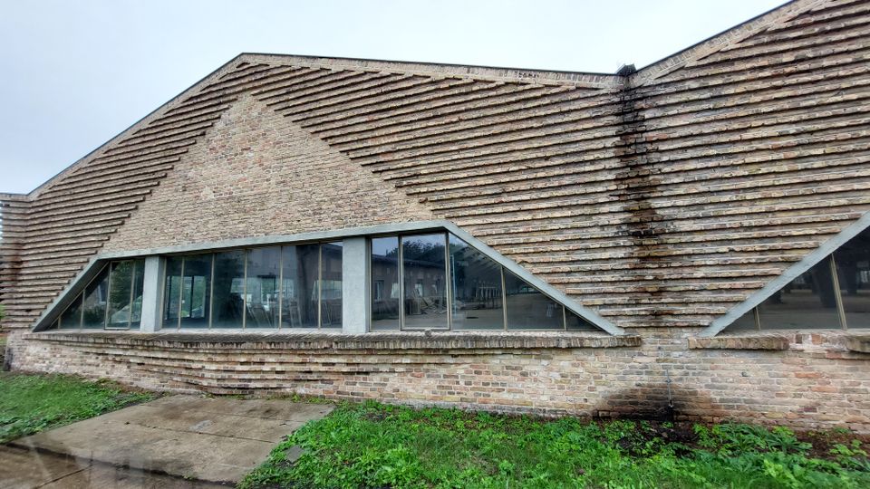 Ein modernes Gebäude aus Ziegelsteinen mit schräg verlaufendem Mauerwerk und großen, trapezförmigen Glasfenstern. Vor dem Gebäude liegt eine grasbedeckte Fläche und ein betonierter Gehweg.