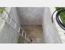 Blick in eine rechteckige, leere Betonbaugrube mit glatten Wänden und Betonboden, in die eine Metallleiter schräg hinabführt. Am oberen Rand ist Gras zu sehen, daneben liegen mehrere aufgerollte schwarze Kunststoffschläuche.