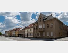 Straßenszene mit mehreren Reihenhäusern im historischen Baustil, teils renoviert, teils sanierungsbedürftig, mit roten und grauen Ziegeldächern. Asphaltierte Straße mit Bürgersteig, Laternen und einer Bushaltestelle; im Hintergrund blauer Himmel mit Wolken.