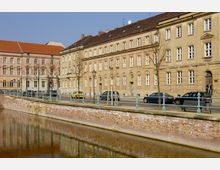 Straßenszene mit historischen, mehrstöckigen Gebäuden aus hellem Putz an einer gepflasterten Straße mit parkenden Autos. Im Vordergrund fließt ein Kanal mit einer niedrigen Ziegelmauer und einer schmiedeeisernen Geländerverzierung.
