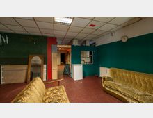 Ein Raum mit grün gestrichenen Wänden, einer weißen Decke mit Rasterleuchten und einem rotbraunen Bodenbelag. Im Vordergrund stehen zwei vintage-gemusterte Sofas und ein Spiegel lehnt an der Wand. Im Hintergrund befinden sich ein kleiner Kühlschrank, ein Wanddurchbruch mit Theke sowie Türen, die in andere Räume führen.