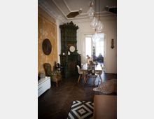 Wohnzimmer mit einem grünen, verzierten Kachelofen als zentralem Element, einem langen Esstisch mit Stühlen im skandinavischen Stil und einem Parkettboden in Fischgrätmuster. An der Wand ist eine historische Putzstruktur zu sehen, ergänzt durch moderne Dekoration, wie hängende Glühbirnen und gemusterte Teppiche.