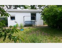 Weißes Gartenhaus mit Flachdach in einem grünen Garten, umgeben von Bäumen und Sträuchern. Davor steht eine Schaukelbank, neben der Pflanzen und Gartenutensilien zu sehen sind.