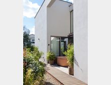 Moderne Hausfassade mit weißem Putz, einer bodentiefen Glastür und einem kleinen Terrassenbereich. Im Vordergrund befinden sich Topfpflanzen, ein runder brauner Whirlpool und ein schmaler Holzsteg, der durch einen Garten mit grüner Bepflanzung führt.