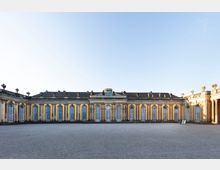 Breiter Gebäudeflügel eines historischen, gelb gestrichenen Schlosses mit symmetrischen, hohen Rundbogenfenstern und dekorativen Statuen auf dem Dachgesims; im Vordergrund ein leerer Kiesplatz unter blauem Himmel.