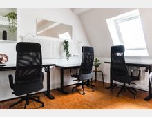 Modernes Büro mit drei schwarzen Bürostühlen an höhenverstellbaren Schreibtischen. Helle Wände, ein schräges Dachfenster, eine Pinnwand mit Pflanzenornamenten und Holzfußboden definieren den Raum.