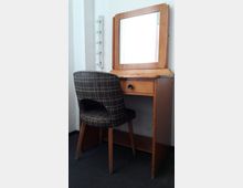 Ein hölzerner Schminktisch mit rechteckigem Spiegel und einer Schublade, daneben eine Wandhalterung mit Glühbirnen. Vor dem Tisch steht ein gepolsterter Stuhl mit kariertem Muster auf schwarzem Teppichboden.