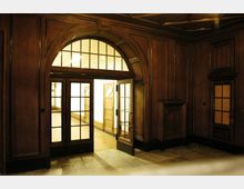 Holzvertäfelter Innenraum mit einer großen, doppelflügeligen Glastür, die von einem hohen Bogen mit Glasfenstern darüber umrahmt wird. Im Hintergrund befindet sich ein heller Flur mit weiteren Holztüren.