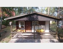 Kleines Ferienhaus mit Holzdach und überdachter Veranda, ausgestattet mit einem Tisch und Stühlen mit gelben Tischdecken. Das Haus liegt in einer bewaldeten Umgebung, mit Bäumen und weiterem Gebäude im Hintergrund.