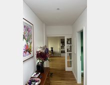 Flur in einem modernen Wohnraum mit Holzboden, weißen Wänden und einer Holzkommode links, dekoriert mit einer Vase voller Blumen. Im Hintergrund ein Wohnzimmer mit Regalen und Glastüren, die den Raum abteilen.