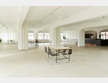 Weitläufiger, moderner Innenraum mit heller Einrichtung, großen Fenstern und weißen Wänden. Zentral ein Tisch mit vier Stühlen, im Hintergrund mehrere Sitzbereiche, ein Bartresen sowie ein vertikales Regal; einige grüne Pflanzen sind sichtbar.